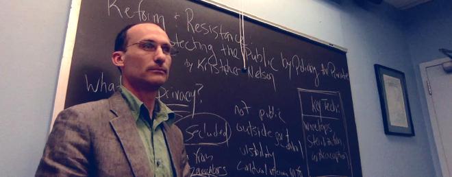 Kristopher standing in front of a chalkboard and looking serious &amp; teacherly.